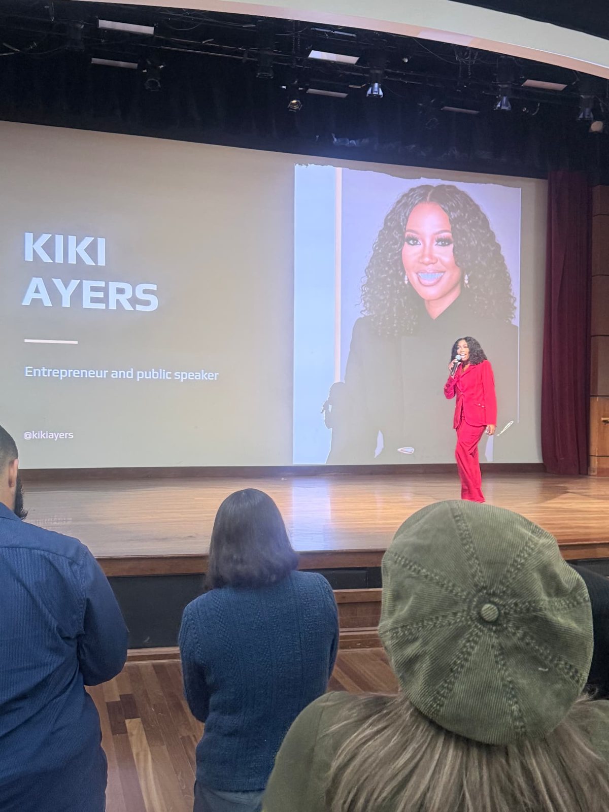 Kiki Ayers on stage in a red suit, her name and photo on the screen behind her